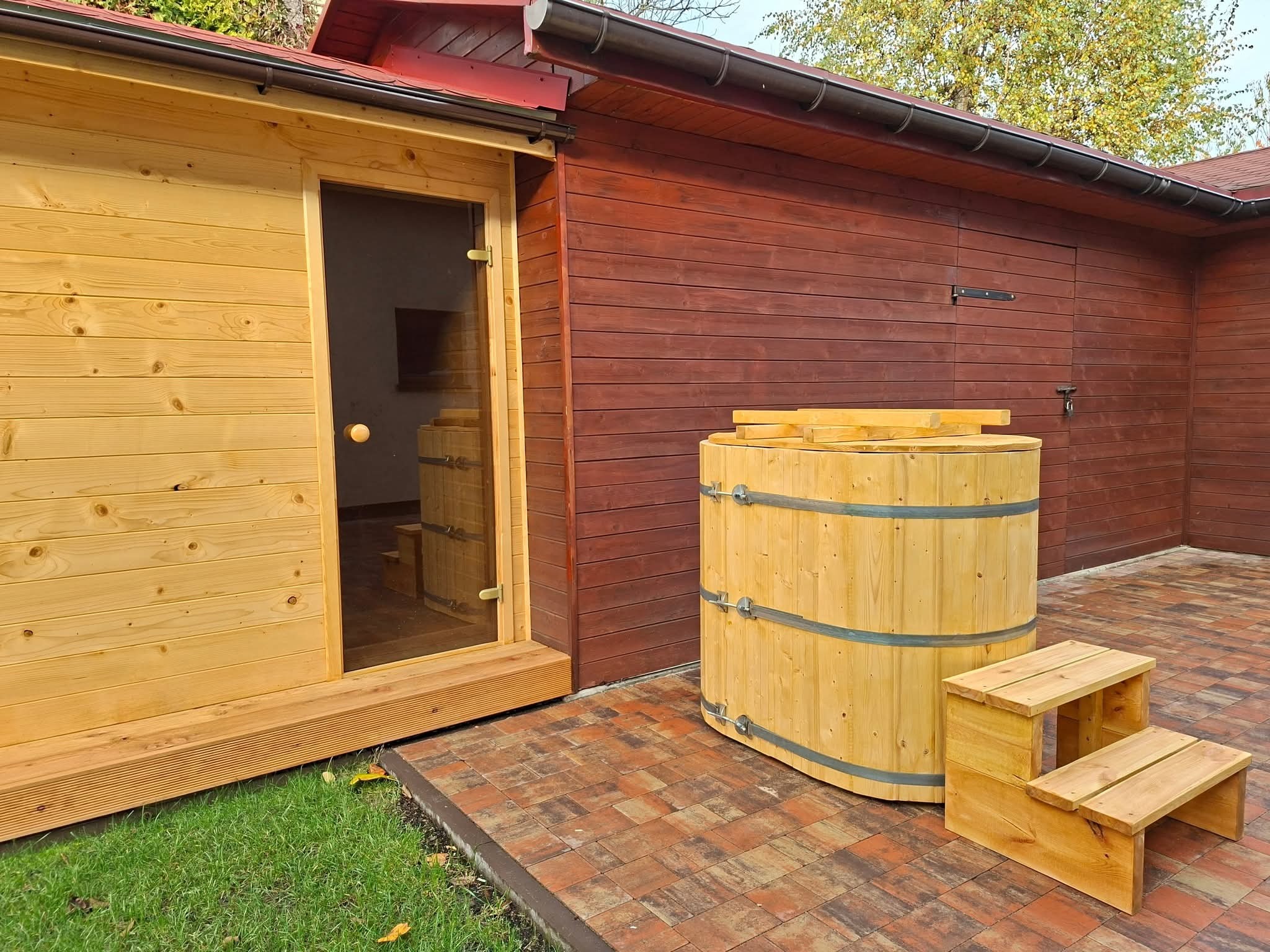 Drewniana sauna z przeszklonymi drzwiami, obok beczka z wodą i schodki. Całość na kostce brukowej, obok trawnik. Budynek z brązowych desek w tle.