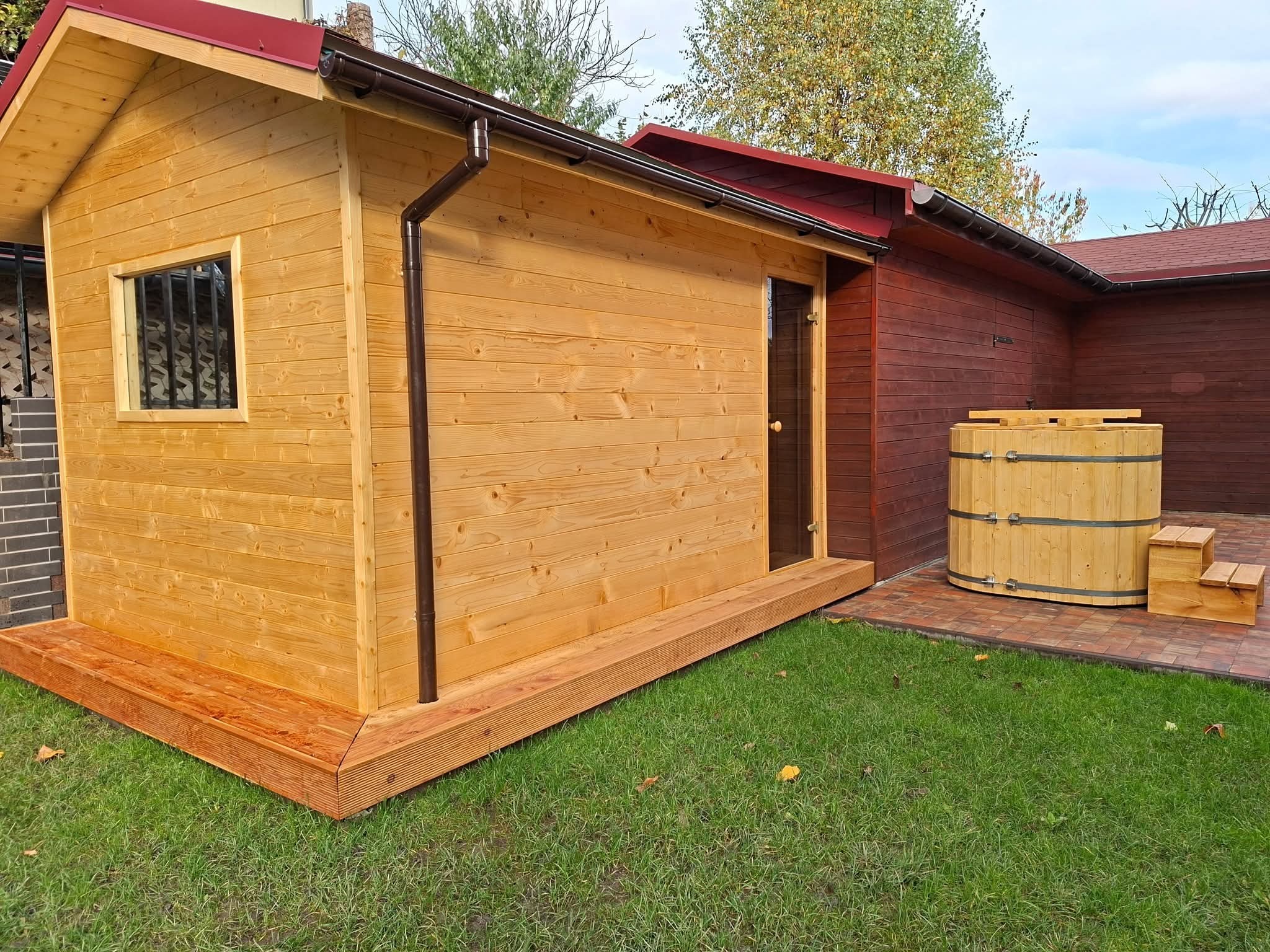 Drewniana sauna ogrodowa z oknem i beczką do schładzania, ustawiona na trawniku. Widoczne brązowe rynny i czerwony dach. Ujęcie z poziomu trawy.