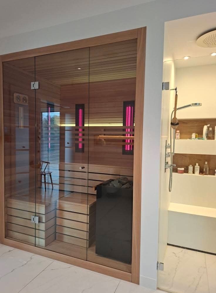 Nowoczesna sauna z przeszklonymi drzwiami i drewnianym wnętrzem, widoczna z perspektywy łazienki z prysznicem. Minimalistyczny design, jasne kolory i subtelne oświetlenie.