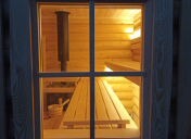 Wnętrze drewnianej kabiny widziane przez okno. W środku piec, ławki i wiaderko z chochlą, całość oświetlona ciepłym światłem.