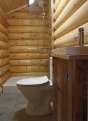 Wnętrze łazienki w domku z bali, widoczna toaleta, umywalka z szafką z ciemnego drewna oraz prysznic, oświetlenie górne, szare płytki podłogowe.