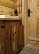 Wnętrze wykończone drewnem: fragment kabiny z drewnianą szafką pod umywalką i drzwiami z czarnym uchwytem, widoczne połączenia bali.