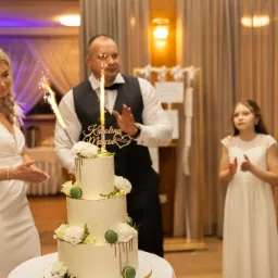 Trzy poziomowy tort weselny dekorowany białymi kwiatami i zielonymi makaronikami, z płonącymi zimnymi ogniami, w tle para młoda i dziewczynka w białych sukienkach