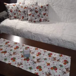 Dekoracyjny bieżnik i poduszki z motywem świątecznym reniferów i prezentów na białej, futrzanej sofie. Bieżnik ułożony na ciemnym, drewnianym stole.