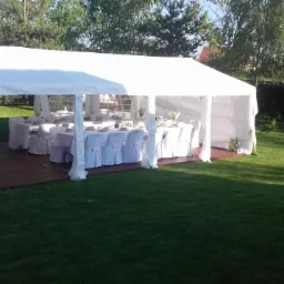 Biały namiot cateringowy na drewnianym podeście, ustawiony na trawniku, z nakrytymi stołami i krzesłami z różowymi kokardkami, przygotowany na imprezę plenerową.