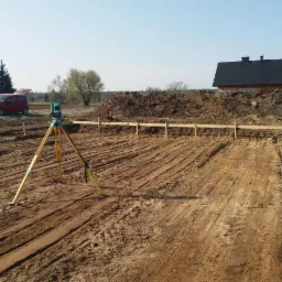 Prace przygotowawcze pod budowę domu: wytyczone fundamenty, drewniane szalunki, niwelacja terenu, teodolit na statywie, czerwony samochód w tle.