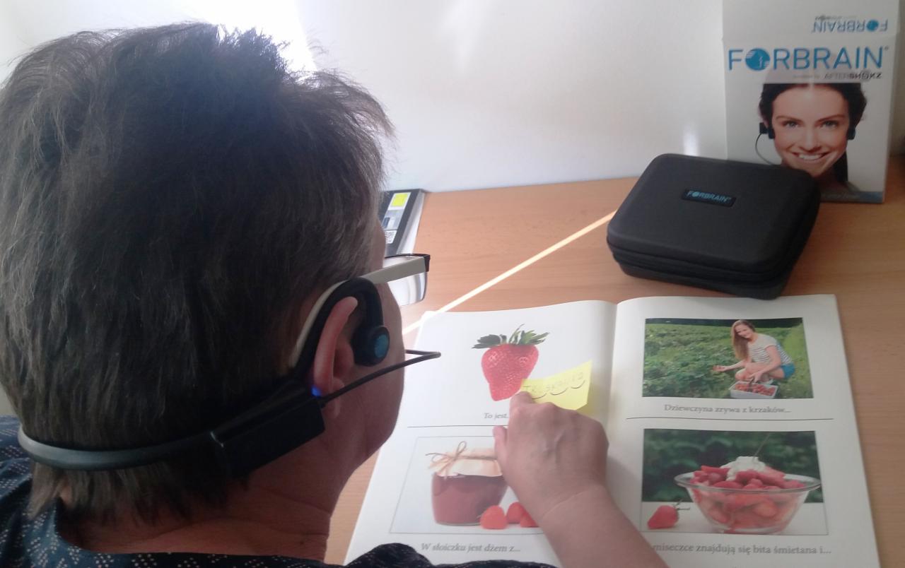 Osoba z założonym urządzeniem Forbrain czyta książkę z obrazkami truskawek, na stole pudełko i opakowanie urządzenia.