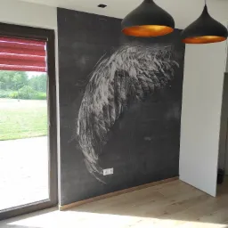 Ściana w pokoju z dekoracyjnym rysunkiem skrzydła, oświetlona przez dwie czarne lampy wiszące z złotym wnętrzem, widok na zewnątrz przez okno z czerwoną roletą.