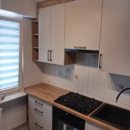 Jasnoszara zabudowa kuchenna z drewnianym blatem i czarnymi uchwytami, widok na płytę gazową i czarny zlew, w tle białe płytki z delikatnym wzorem i okno z żaluzjami poziomymi.