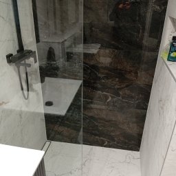 Usługi Remontowe - Nowoczesna kabina prysznicowa z ciemnymi płytkami imitującymi marmur i armaturą w kolorze czarnym. Widoczny odpływ liniowy i fragment szafki umywalkowej.