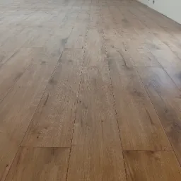 Ukończona podłoga z paneli imitujących drewno w jasnym odcieniu brązu, ułożona w nowym pomieszczeniu z białymi ścianami i widocznym gniazdkiem elektrycznym.