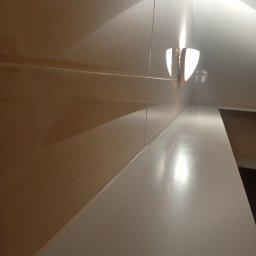 Usługi Remontowe - Błyszczące beżowe płytki ścienne z poziomymi fugami, biały gładki blat i nowoczesny kinkiet, tworzące odbicia światła.