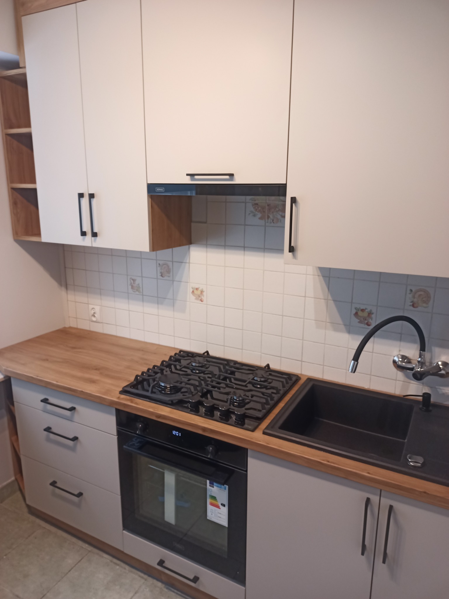 Wykończona kuchnia z jasnymi szafkami górnymi i szarymi dolnymi, drewniany blat, czarny zlew i płyta gazowa, ozdobne płytki ścienne.