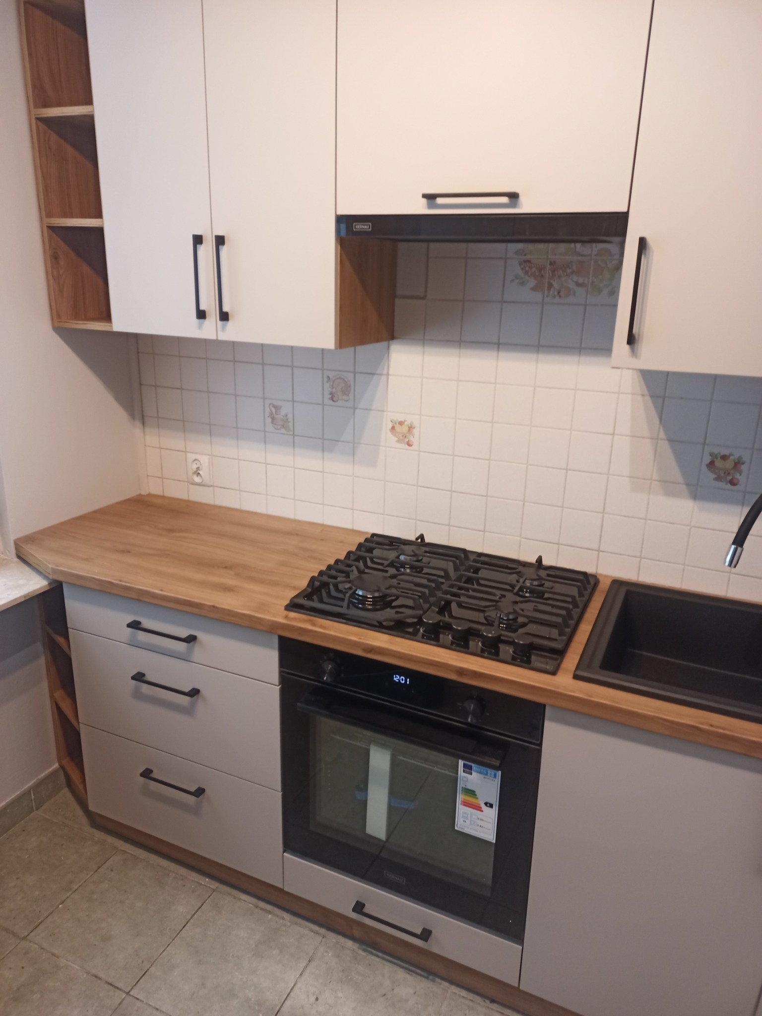 Jasna kuchnia z drewnianym blatem, czarnymi uchwytami i kwadratowymi płytkami z motywami kwiatowymi na ścianie.