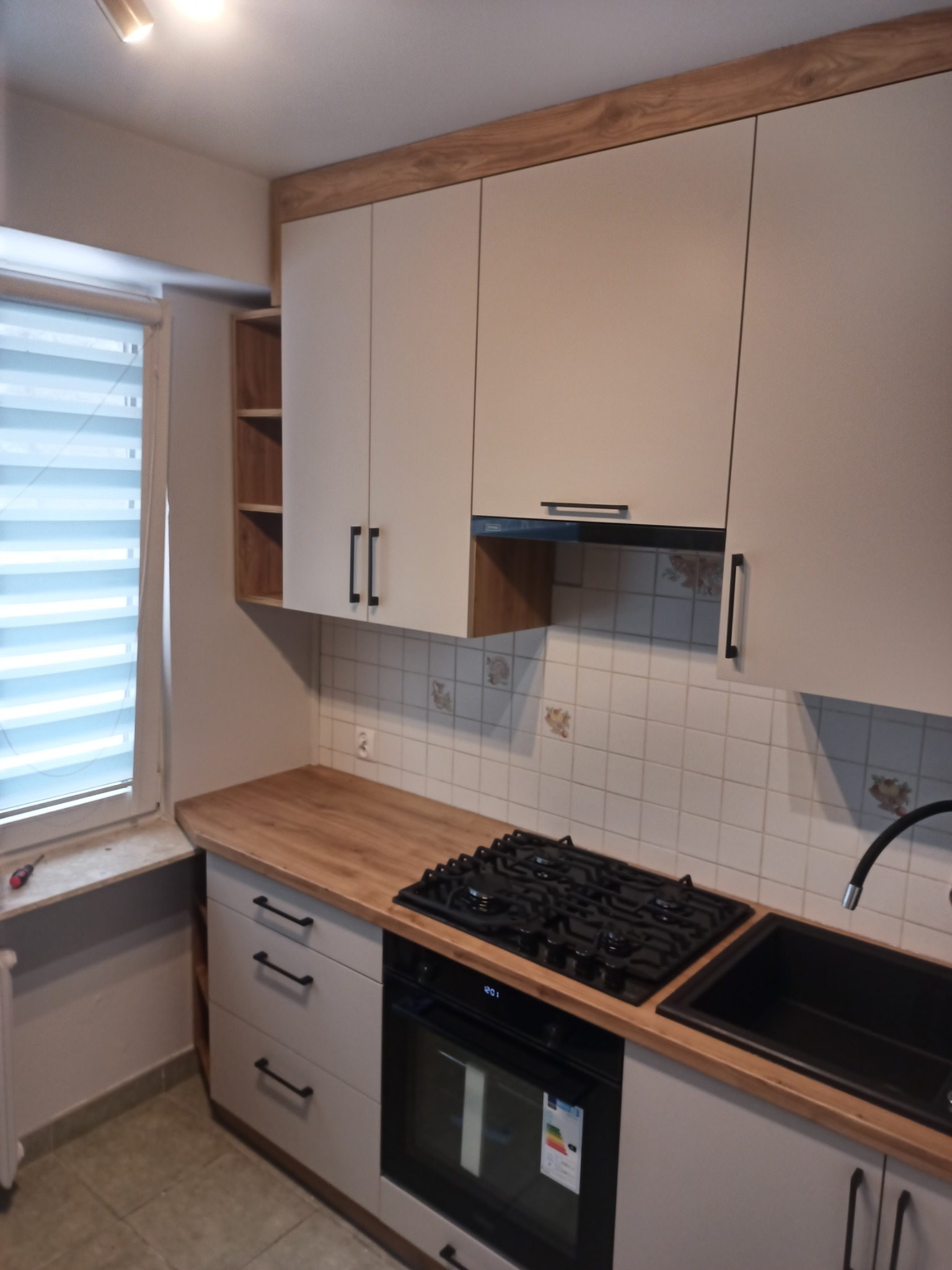 Jasnoszara zabudowa kuchenna z drewnianym blatem i czarnymi uchwytami, widok na płytę gazową i czarny zlew, w tle białe płytki z delikatnym wzorem i okno z żaluzjami poziomymi.