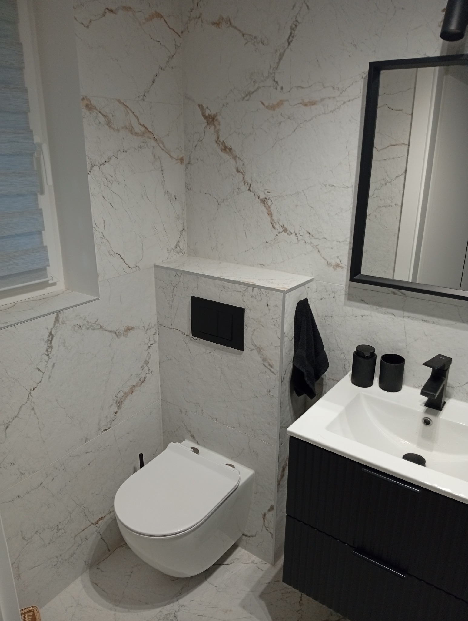 Nowoczesna łazienka z marmurowymi płytkami, podwieszaną toaletą i czarną armaturą. Minimalistyczny design z akcentami w postaci czarnych dodatków.