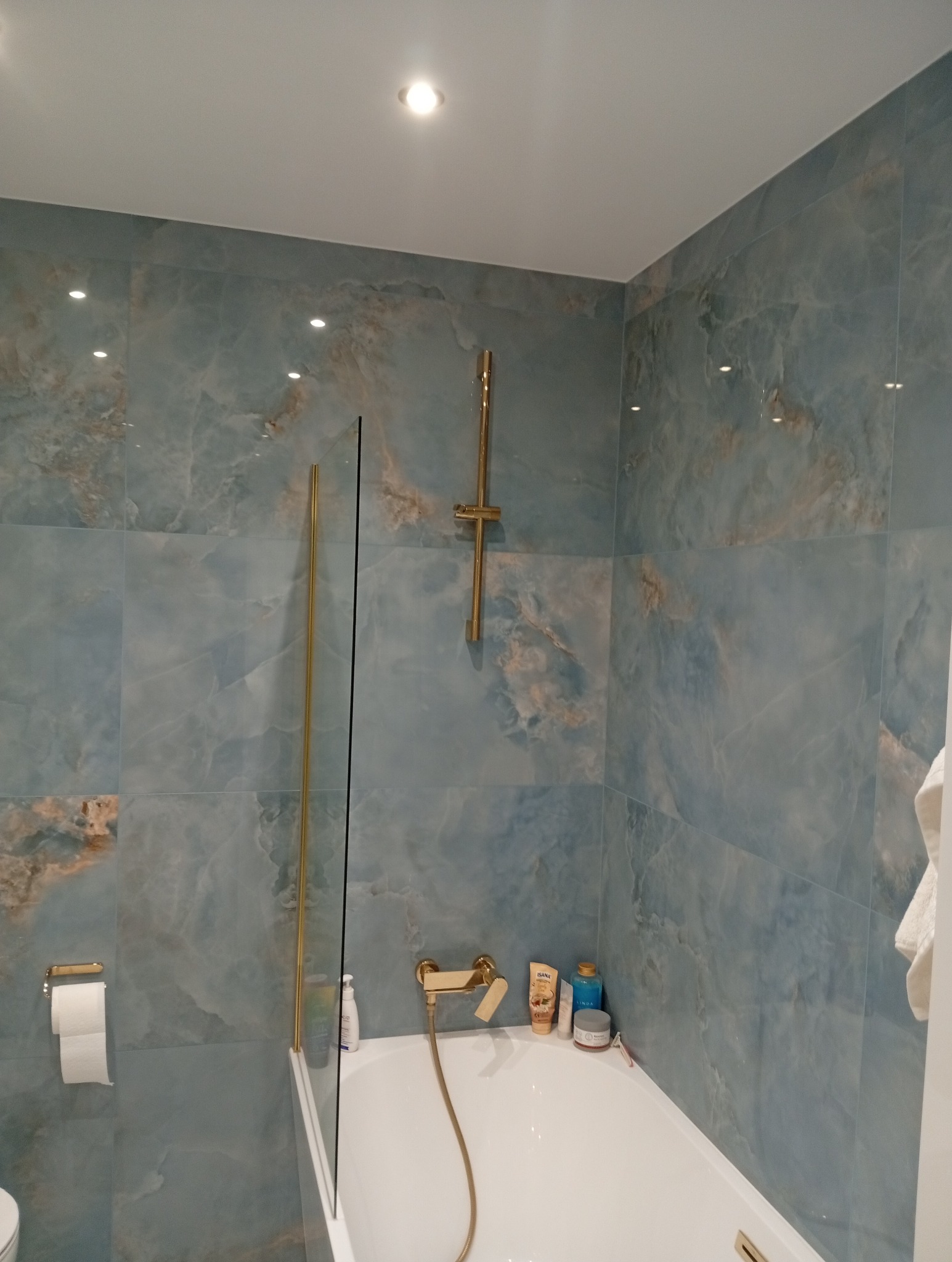 Błękitna łazienka z wanną i złotą armaturą. Płytki imitujące marmur, szklany parawan nawannowy. Nowoczesny design, oświetlenie punktowe.