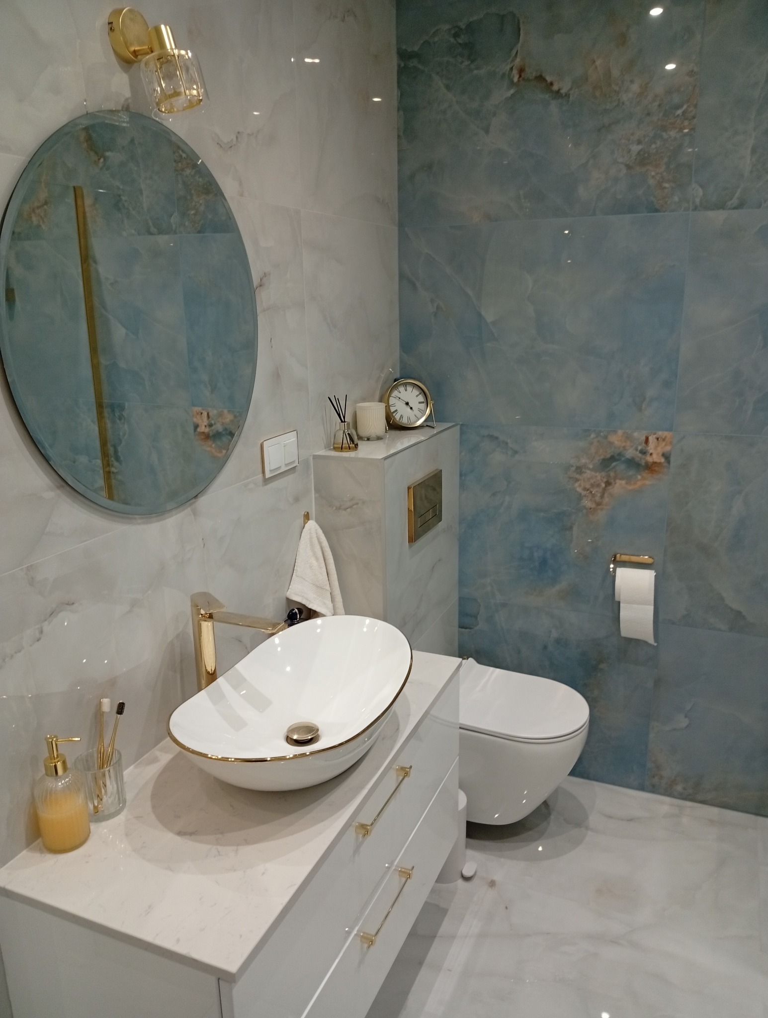 Elegancka łazienka z umywalką nablatową, okrągłym lustrem i płytkami imitującymi marmur w odcieniach błękitu i bieli. Złote akcenty armatury i dodatków.