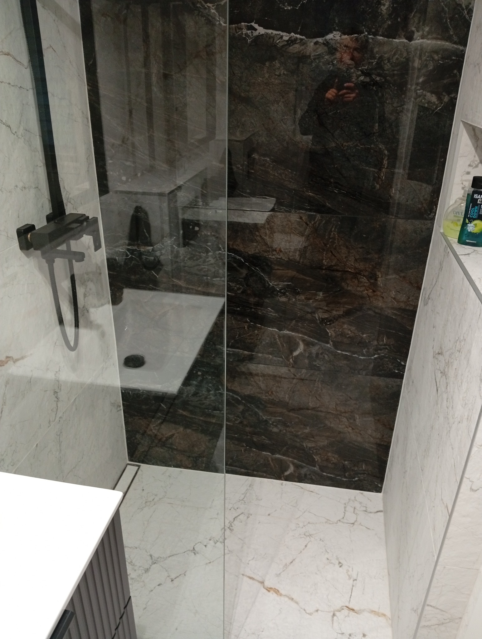 Nowoczesna kabina prysznicowa z ciemnymi płytkami imitującymi marmur i armaturą w kolorze czarnym. Widoczny odpływ liniowy i fragment szafki umywalkowej.