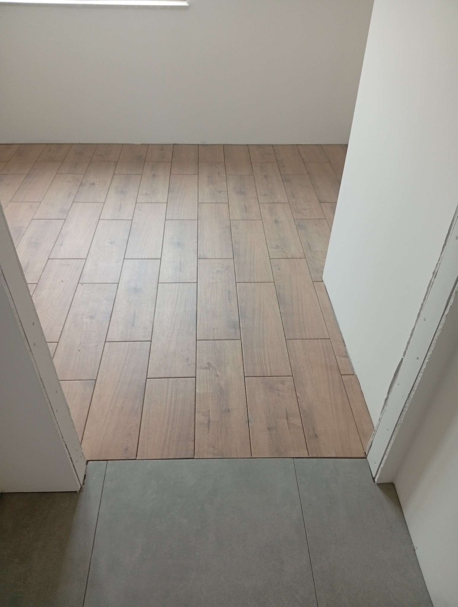 Ukończona podłoga z płytek imitujących drewno, przechodząca w szare płytki przy progu. Widok z góry, neutralne oświetlenie, fragment ściany i ościeżnicy.
