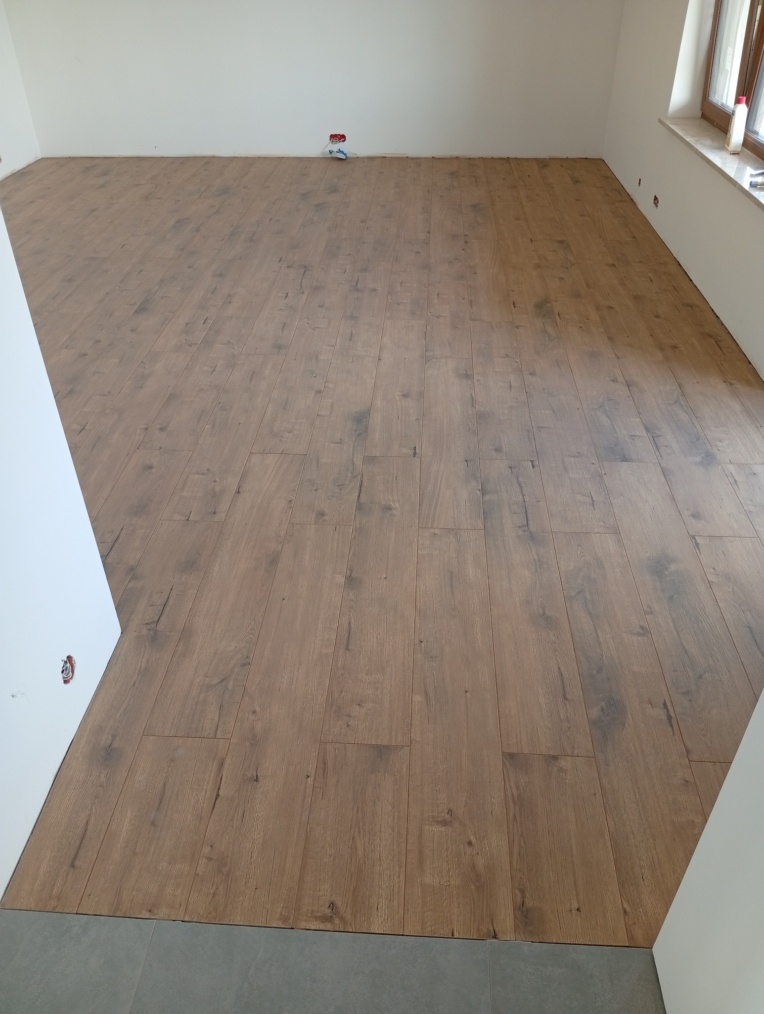 Ułożona podłoga z paneli imitujących drewno w jasnym pomieszczeniu. Widoczne gniazdko elektryczne i fragment szarej podłogi przy ścianie. Ujęcie z niskiego kąta.