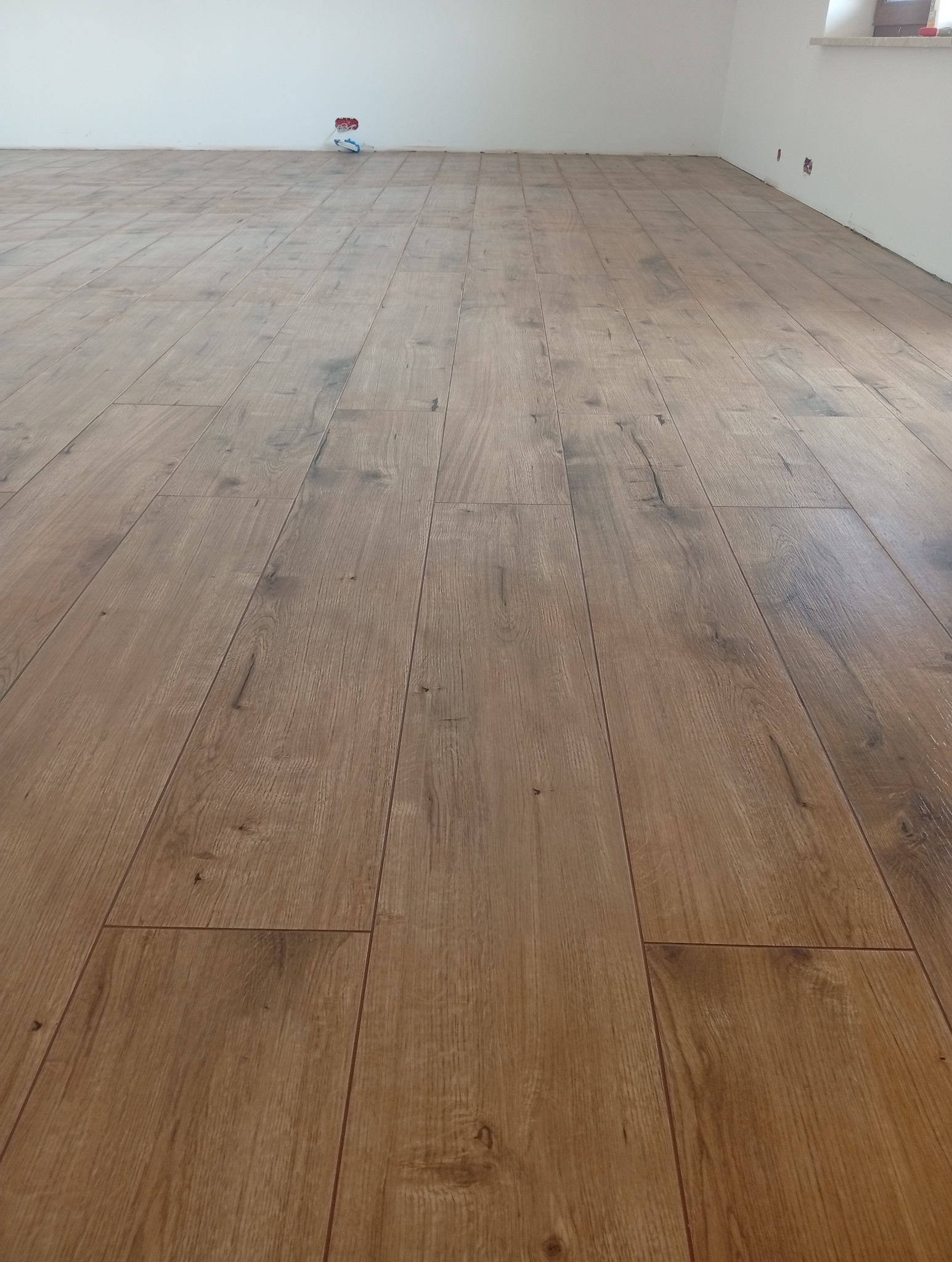 Ukończona podłoga z paneli imitujących drewno w jasnym odcieniu brązu, ułożona w nowym pomieszczeniu z białymi ścianami i widocznym gniazdkiem elektrycznym.