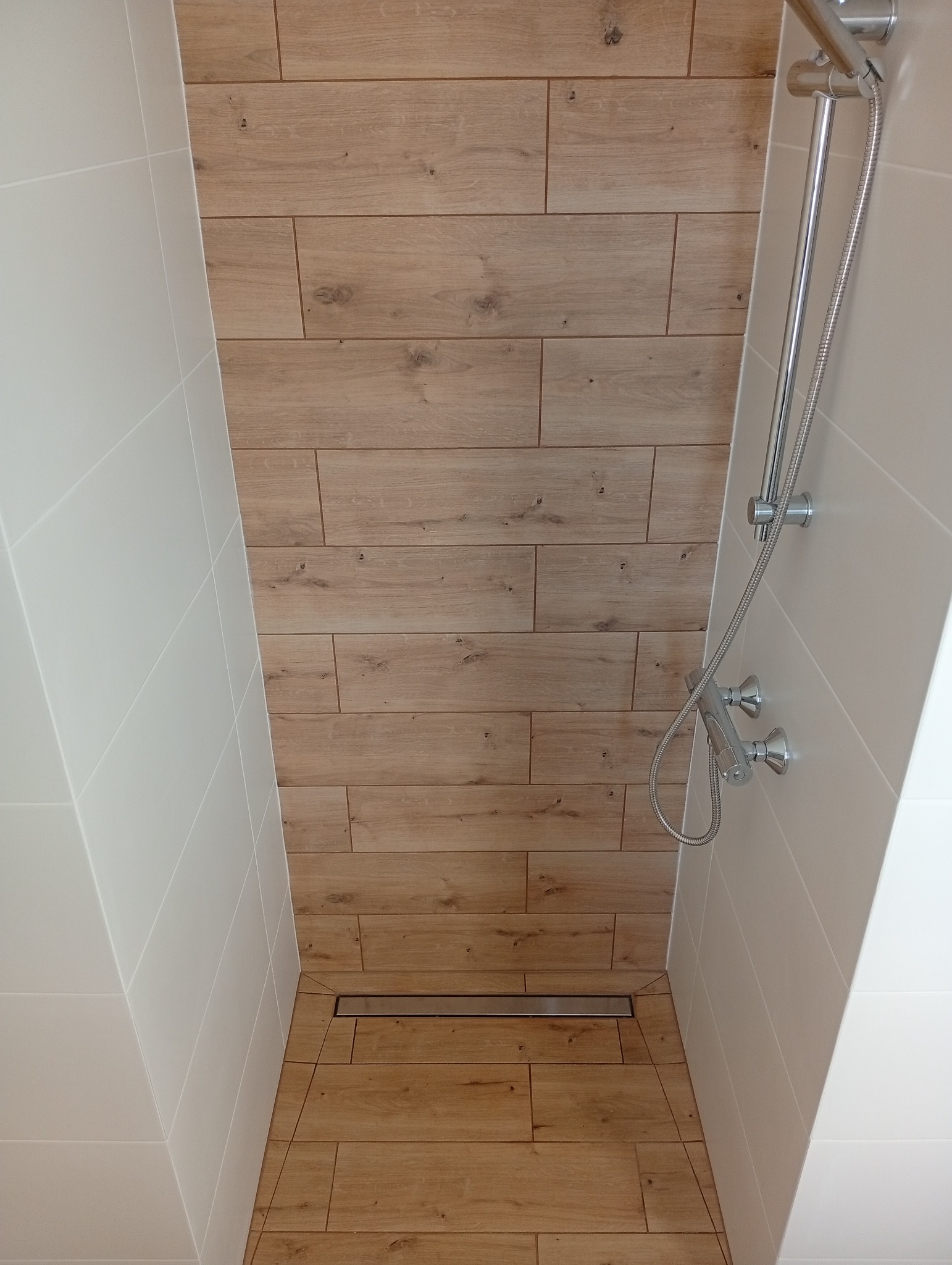 Nowoczesny prysznic walk-in z drewnopodobnymi płytkami i odpływem liniowym. Białe, gładkie płytki na ścianach. Chromowana bateria natryskowa.