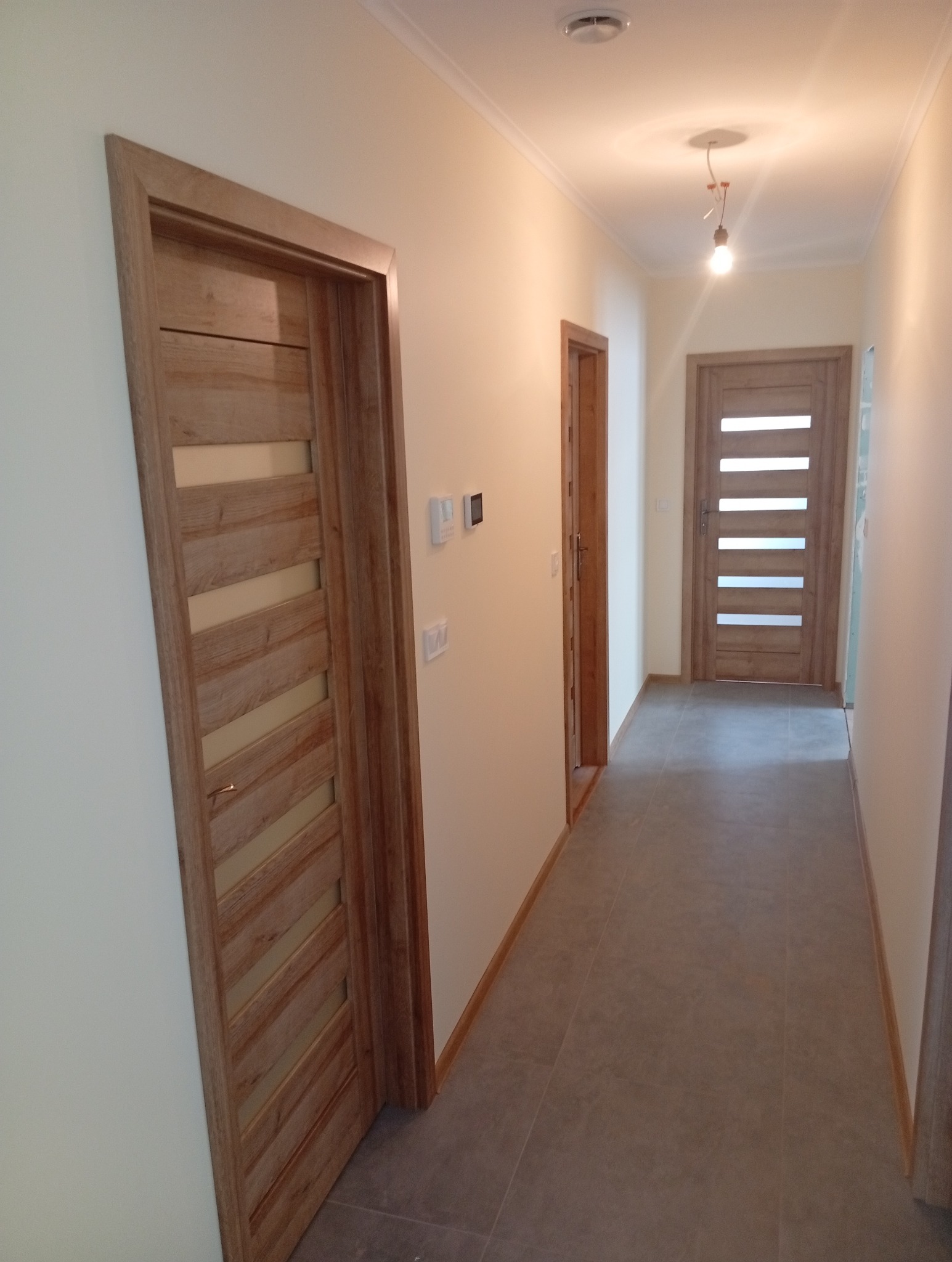 Wykończony korytarz z szarą podłogą i drewnianymi drzwiami z przeszkleniami. Widoczne detale wykończeniowe, takie jak listwy przypodłogowe i oświetlenie sufitowe.