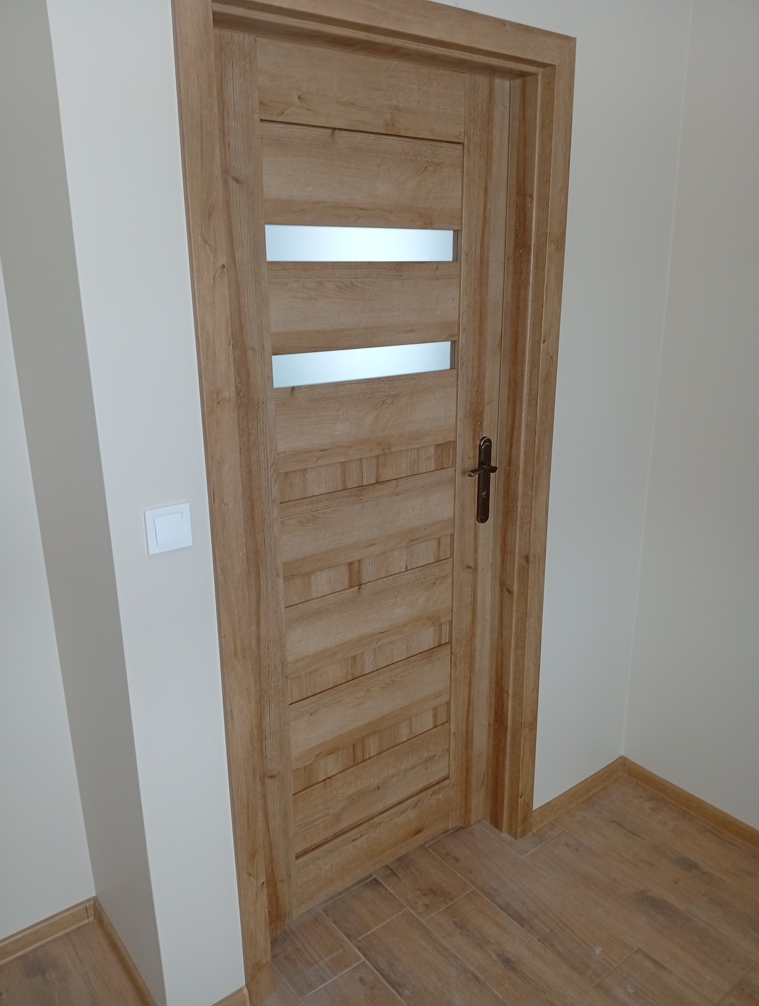 Zamknięte, drewniane drzwi wewnętrzne z trzema poziomymi przeszkleniami, zamontowane w jasnym pomieszczeniu z panelami podłogowymi w odcieniach brązu.