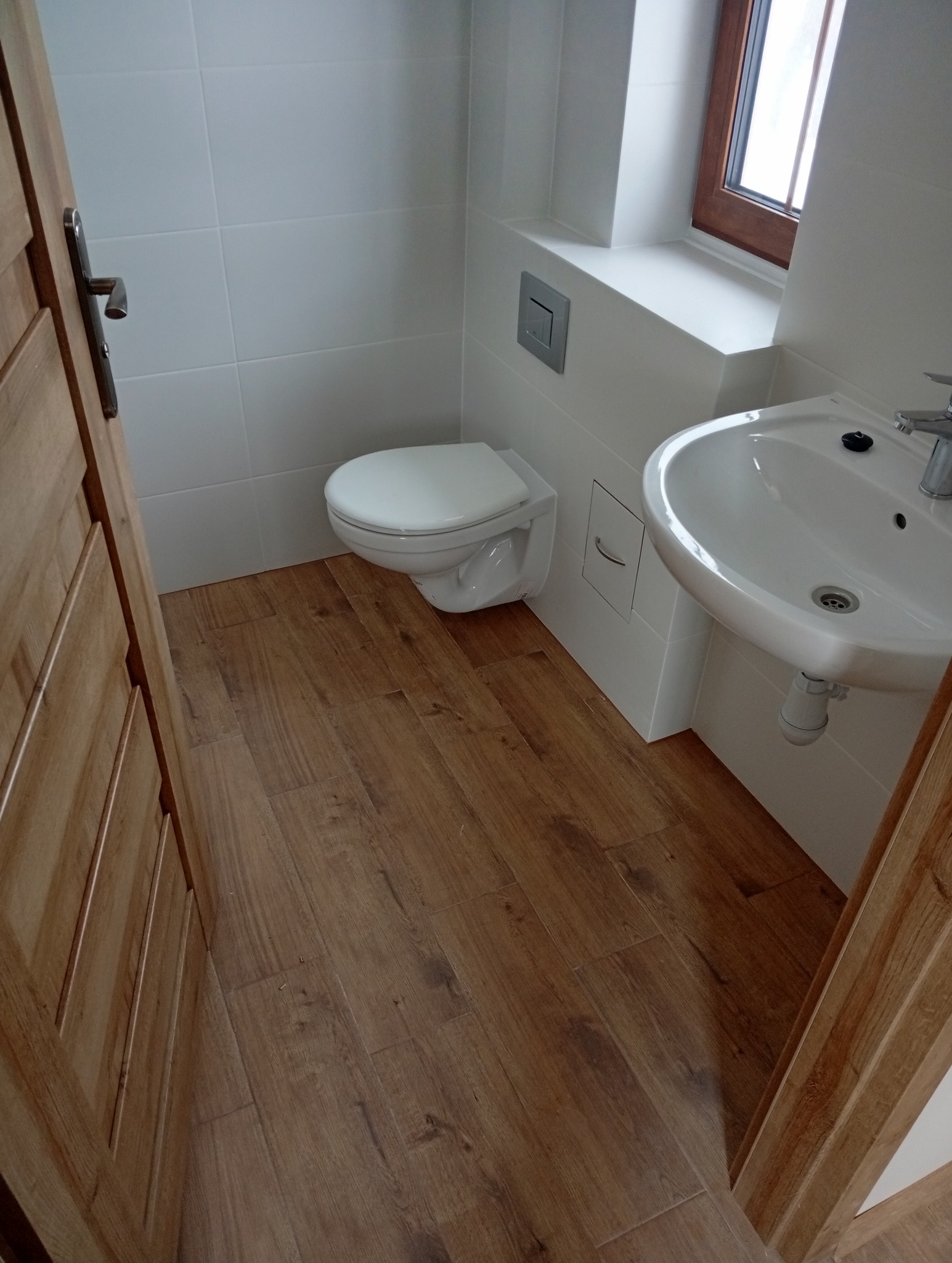 Nowoczesna łazienka po remoncie: białe płytki, podwieszana toaleta, umywalka i drewnopodobna podłoga. Widok na okno i drzwi z drewnianą ramą.