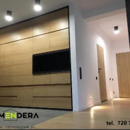 Nowoczesne wnętrze z zabudową meblową z jasnego drewna, czarną ramą i wbudowanym telewizorem, minimalistyczne oświetlenie sufitowe, ściana z dekoracyjną strukturą.