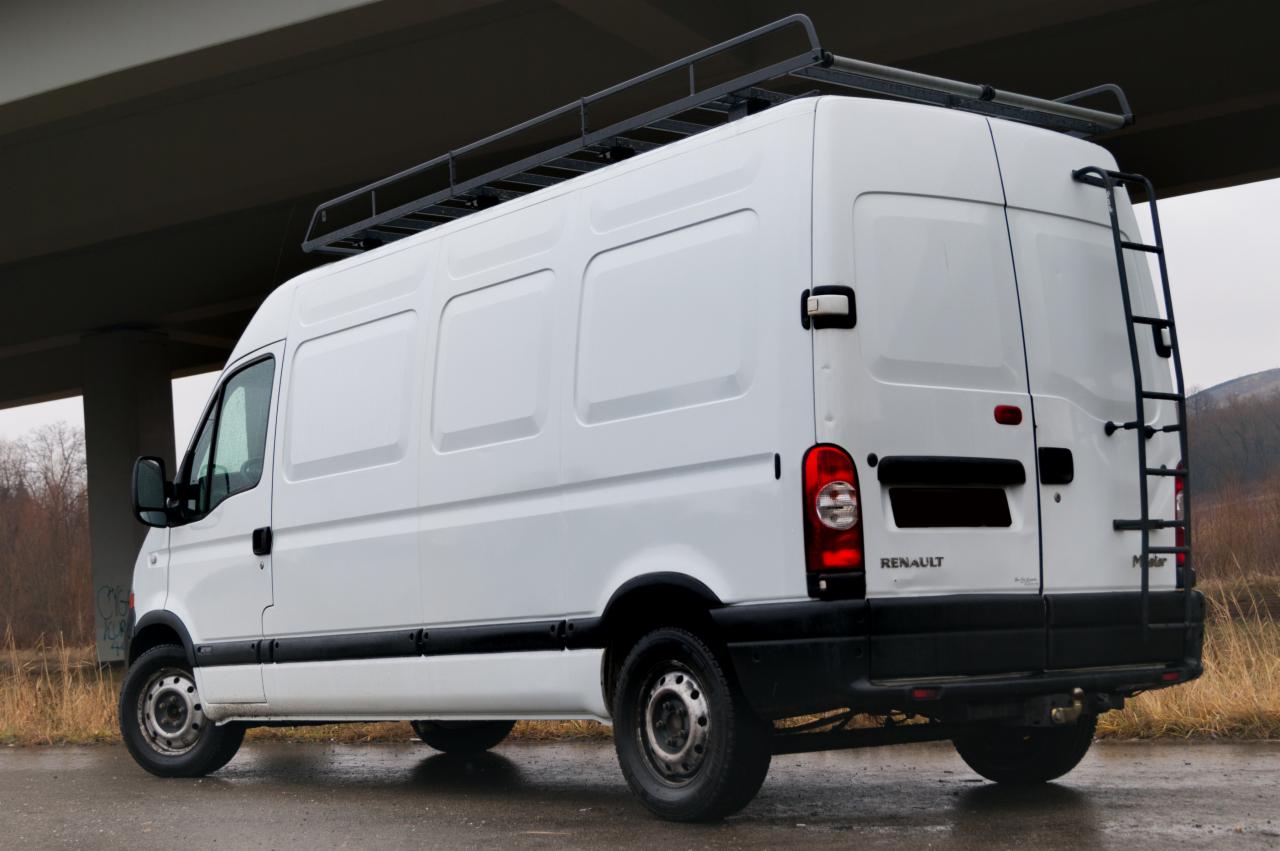 Biały Renault Master z drabinką i bagażnikiem dachowym sfotografowany z tyłu na tle betonowego wiaduktu i łąki.