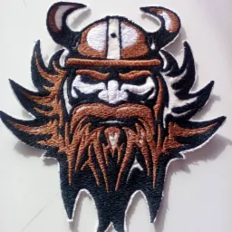 Naszywka z haftowanym wizerunkiem Wikinga w hełmie z rogami, wykonana z czarnej, brązowej i białej nici na białym tle.