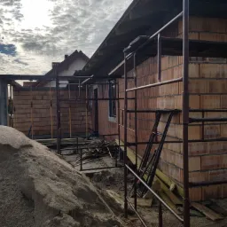 Wznoszenie ścian z bloczków ceramicznych na budowie domu jednorodzinnego, widoczne metalowe rusztowania i sterta piasku.