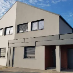 Nowoczesny dom jednorodzinny z szarą elewacją, ciemnymi ramami okiennymi i metalowymi panelami dekoracyjnymi pod okapem, na tle błękitnego nieba z chmurami.