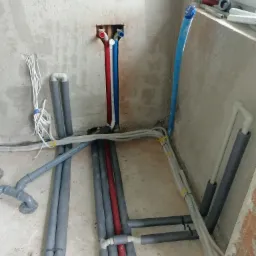 Instalacja wodna i grzewcza w surowym stanie: rury w izolacji szarej i czerwonej, niebieska rura przy oknie, białe kable przy ścianie, widoczne połączenia i przejścia przez ścianę.