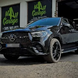 Czarny Mercedes GLE po detailingu, zaparkowany przed warsztatem M AH Garage z widocznym logo i numerem telefonu, w tle auto z folią zmieniającą kolor.