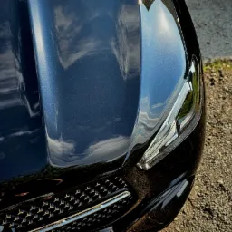 Błyszcząca, granatowa maska samochodu z odbiciem chmur, widoczny reflektor i fragment atrapy chłodnicy.