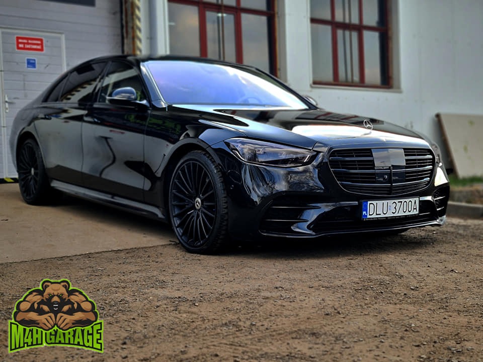 Czarny Mercedes S-klasa z przyciemnionymi szybami, zaparkowany przed garażem, z widocznym logo firmy M4H Garage w lewym dolnym rogu.