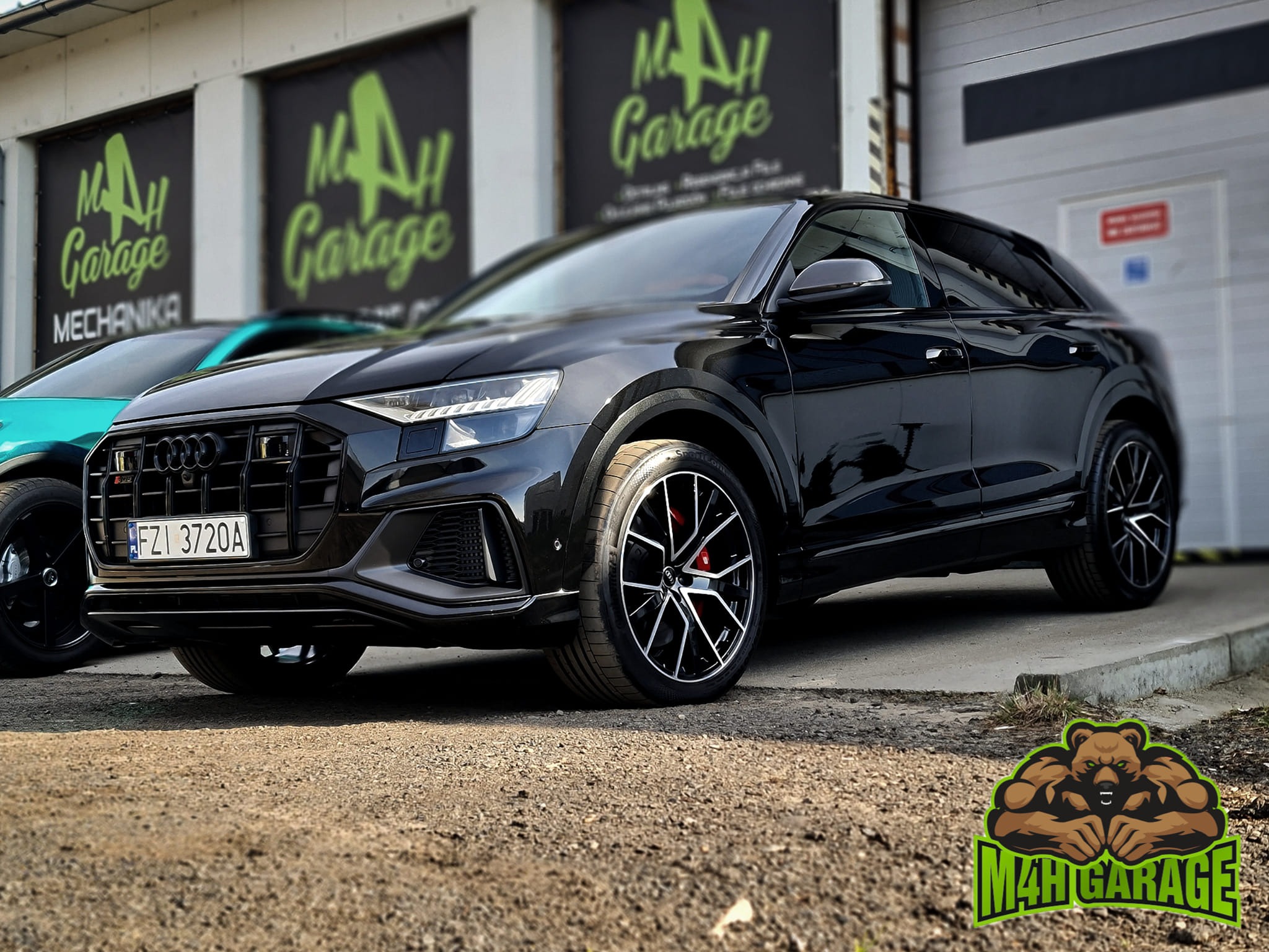 Czarny, luksusowy SUV Audi SQ8 na tle warsztatu M4H Garage w Zielonej Górze. Widoczne logo firmy na drzwiach garażowych i naklejka na podłożu. Charakterystyczne felgi i czerwone zaciski hamulcowe.