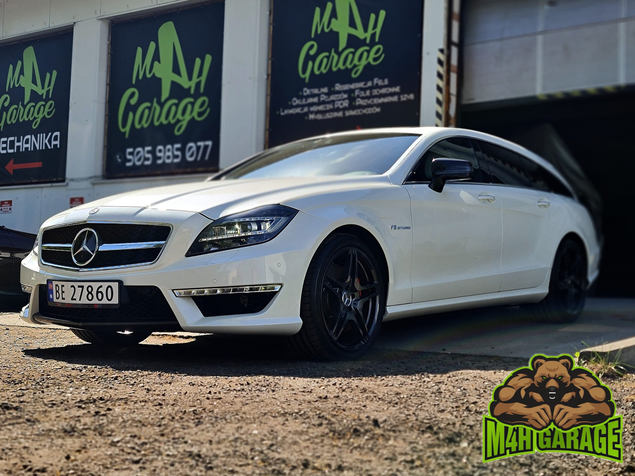 Biały Mercedes-Benz CLS Shooting Brake na tle warsztatu M4H Garage z widocznymi banerami reklamowymi i logo firmy na ziemi.