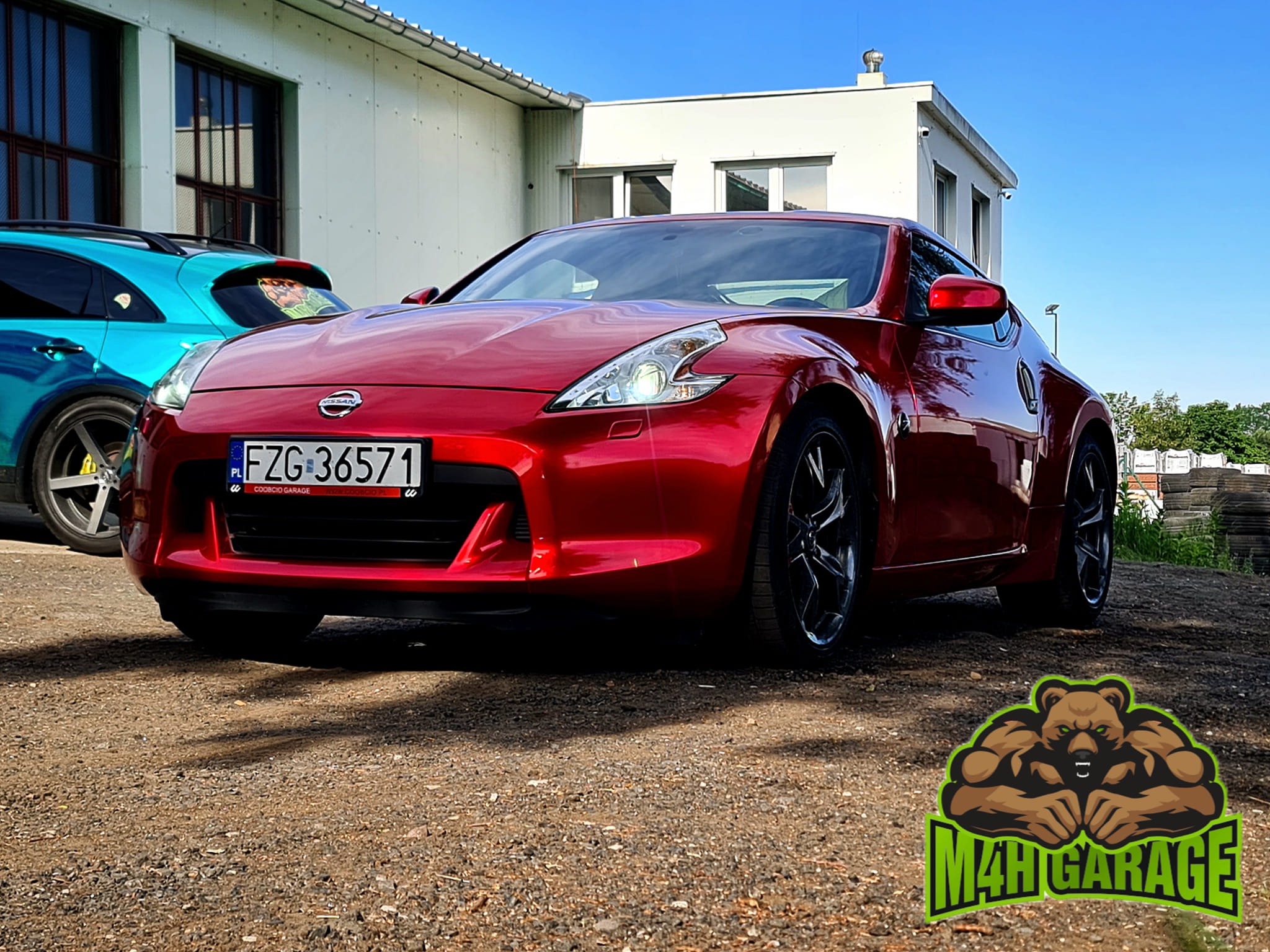 Czerwone sportowe auto Nissan 370Z z czarnymi felgami na żwirowym podłożu przed budynkiem, w tle błękitny samochód i logo firmy M4H Garage w prawym dolnym rogu.