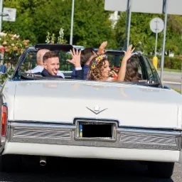 Para młoda w białym kabriolecie Cadillac De Ville z kwiatowym wiankiem na głowie panny młodej, widziana z tyłu, z uniesionymi rękami w geście radości podczas przejazdu przez miasto.