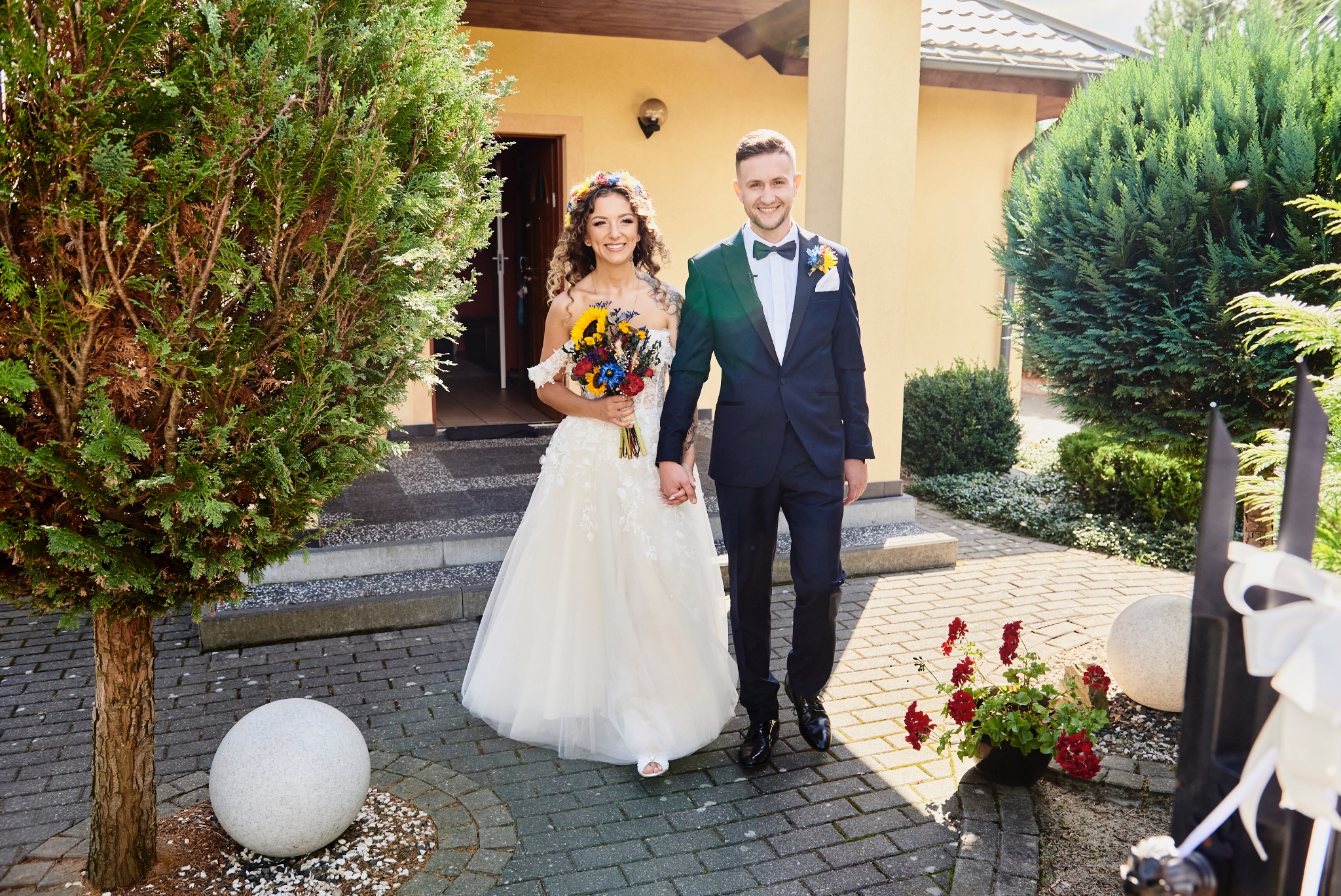 Uśmiechnięta para młoda trzymająca się za ręce, panna młoda w białej sukni z kwiatami we włosach i bukietem polnych kwiatów, pan młody w granatowym garniturze z muszką, na tle budynku z jasną...