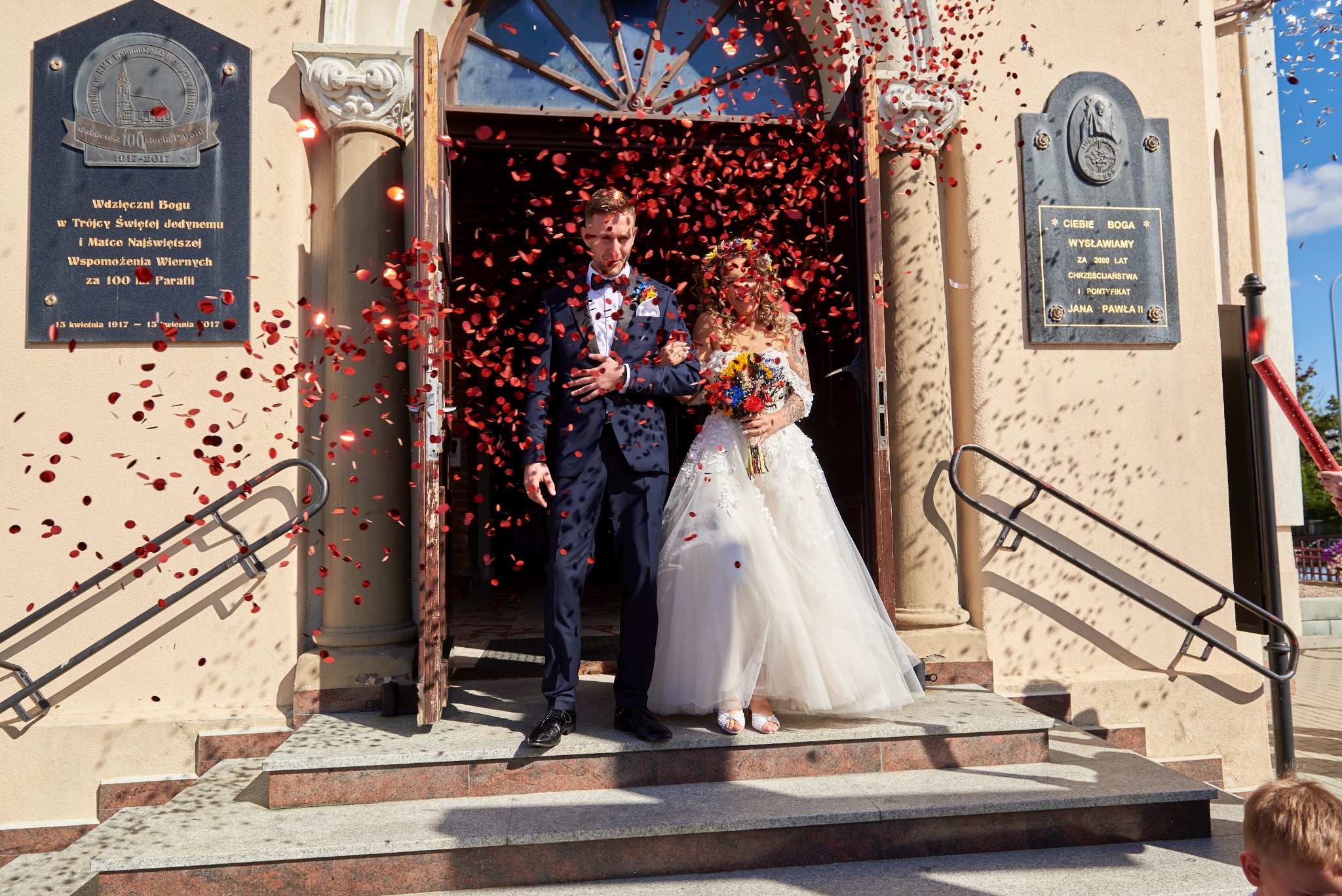 Para młoda wychodzi z kościoła, obsypana czerwonymi płatkami konfetti, panna młoda w białej sukni z bukietem kolorowych kwiatów, pan młody w granatowym garniturze z muszką.