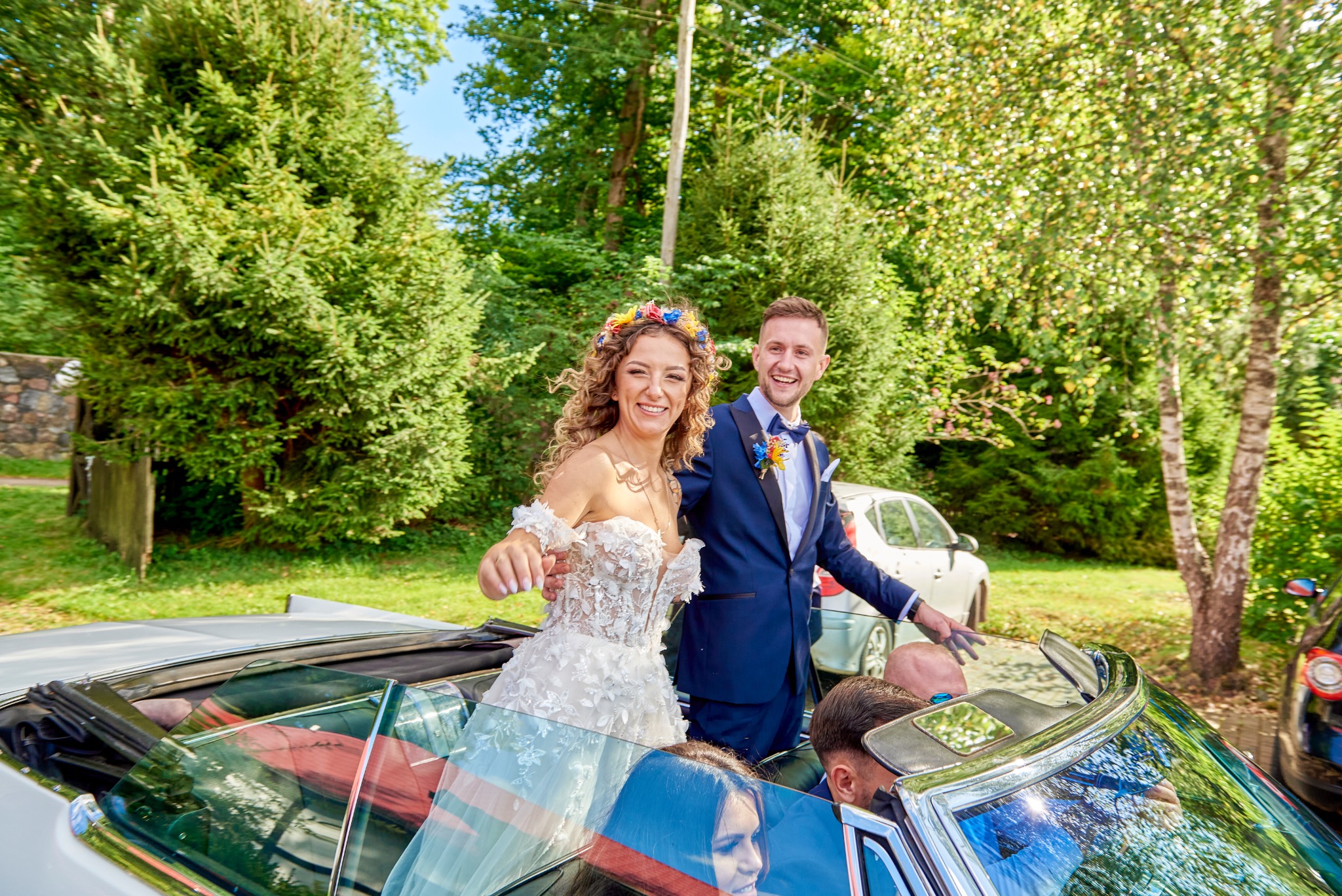 Uśmiechnięta para młoda w kabriolecie z opuszczonym dachem, panna młoda w koronkowej sukni z kwiatowym wiankiem, pan młody w granatowym garniturze z muszką, tło zieleni drzew.