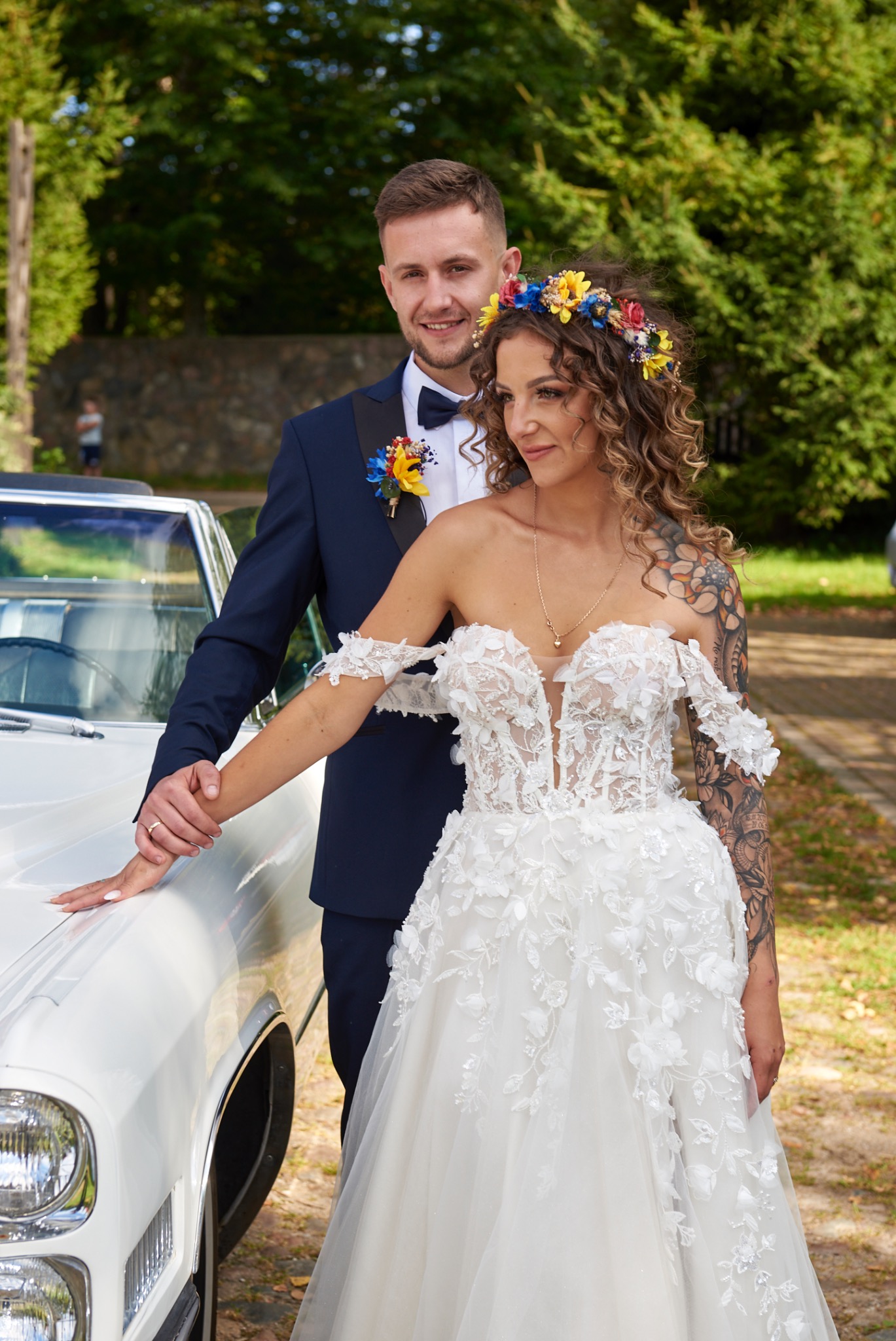 Para młoda pozuje przy białym kabriolecie: panna młoda w koronkowej sukni z odsłoniętymi ramionami i wiankiem z kwiatów, pan młody w granatowym smokingu z butonierką.