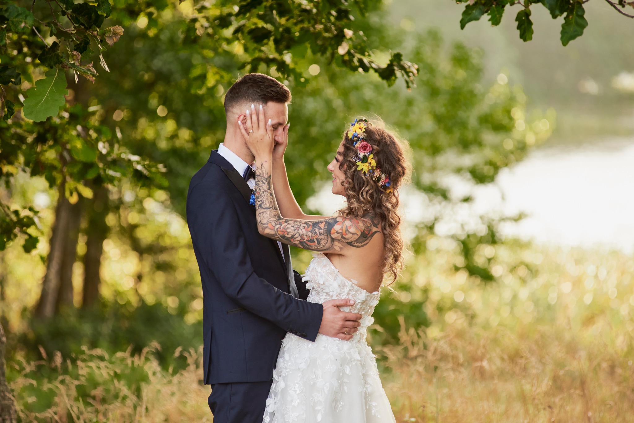 Para młoda w plenerze: Panna młoda z tatuażami i wiankiem na głowie dotyka twarzy pana młodego w eleganckim garniturze, oboje uśmiechnięci na tle zieleni.