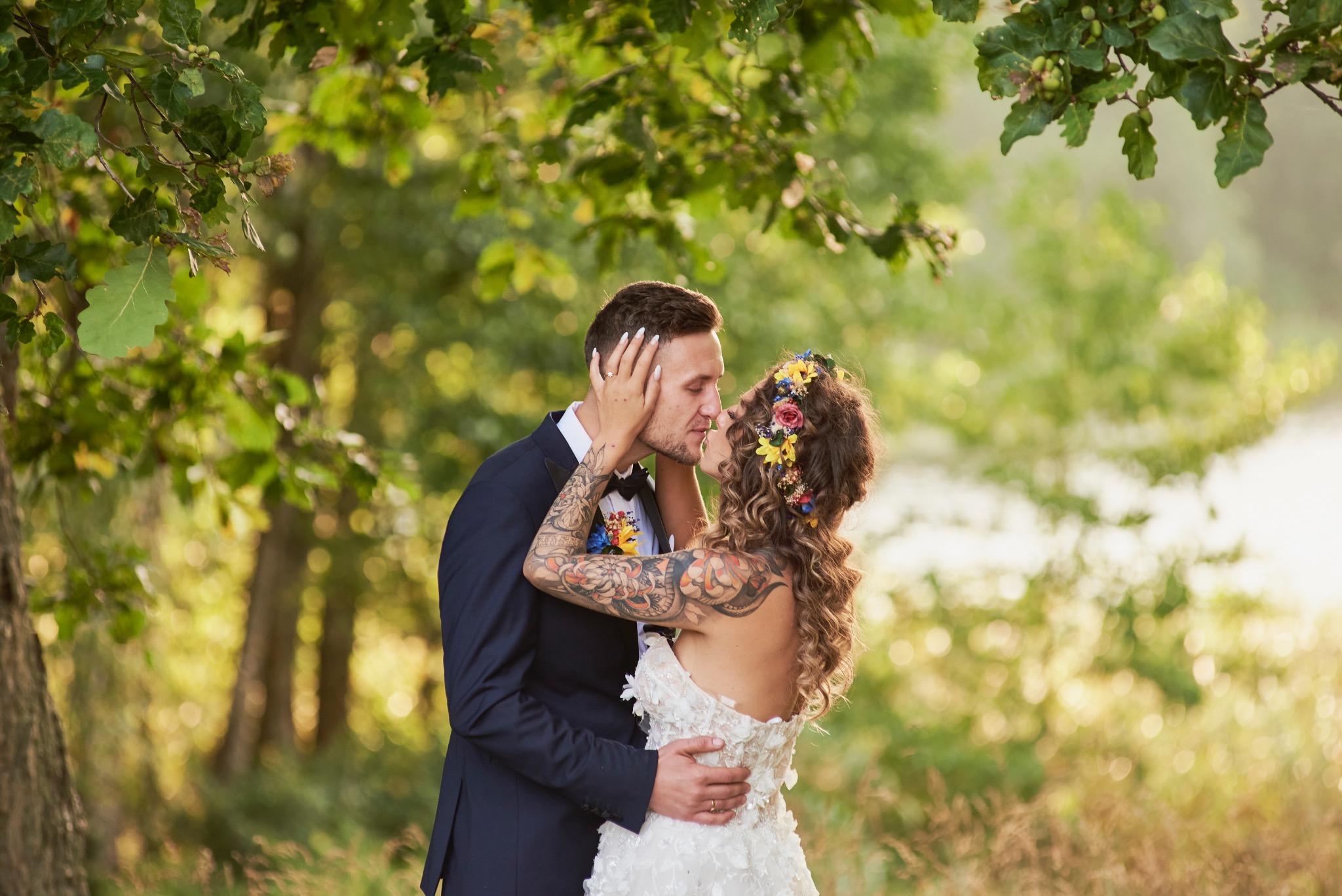 Pocałunek pary młodej w plenerze, panna młoda w sukni z kwiatowymi aplikacjami i wiankiem, pan młody w granatowym garniturze i z tatuażami na rękach, miękkie, rozproszone światło.