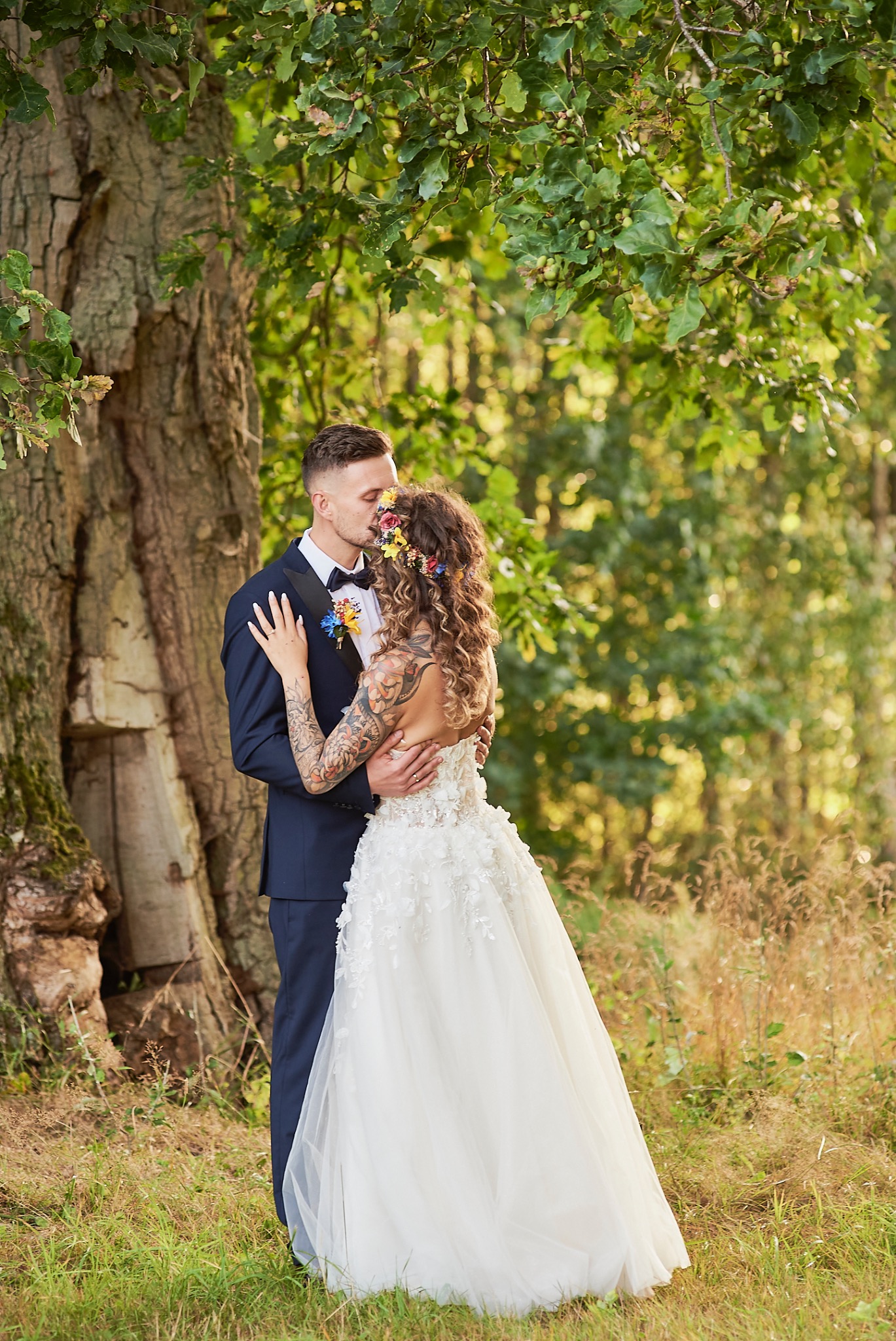 Para młoda w objęciach, panna młoda z kwiatowym wiankiem i tatuażami na ramieniu, w tle duży dąb i leśne otoczenie.